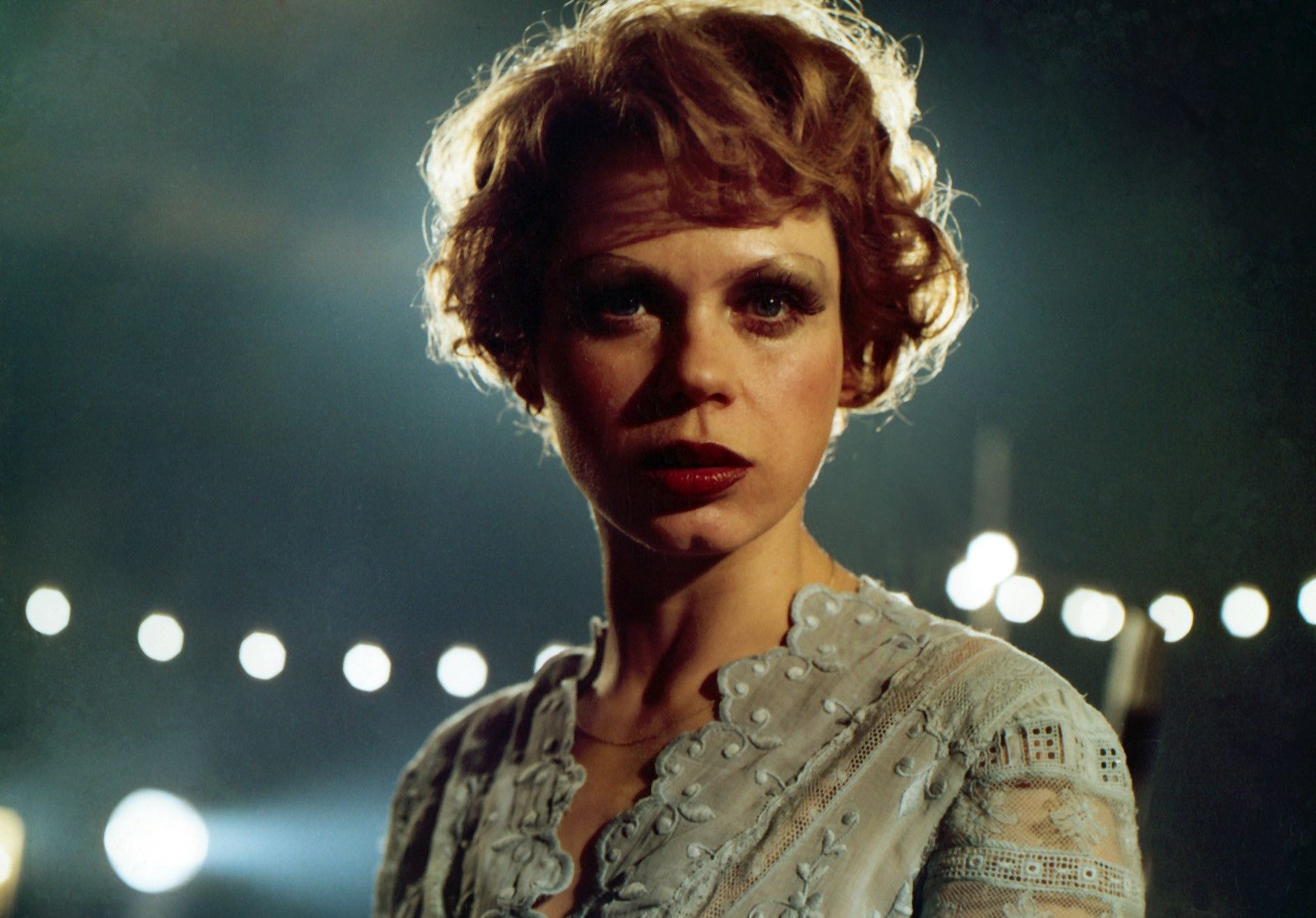 Photo of the actress Renate Krößner as Sunny in the film Solo Sunny (1980). The young woman is wearing a light-coloured embroidered blouse with a floral pattern, red lipstick and short hair. A string of lights can be seen blurred in the background.