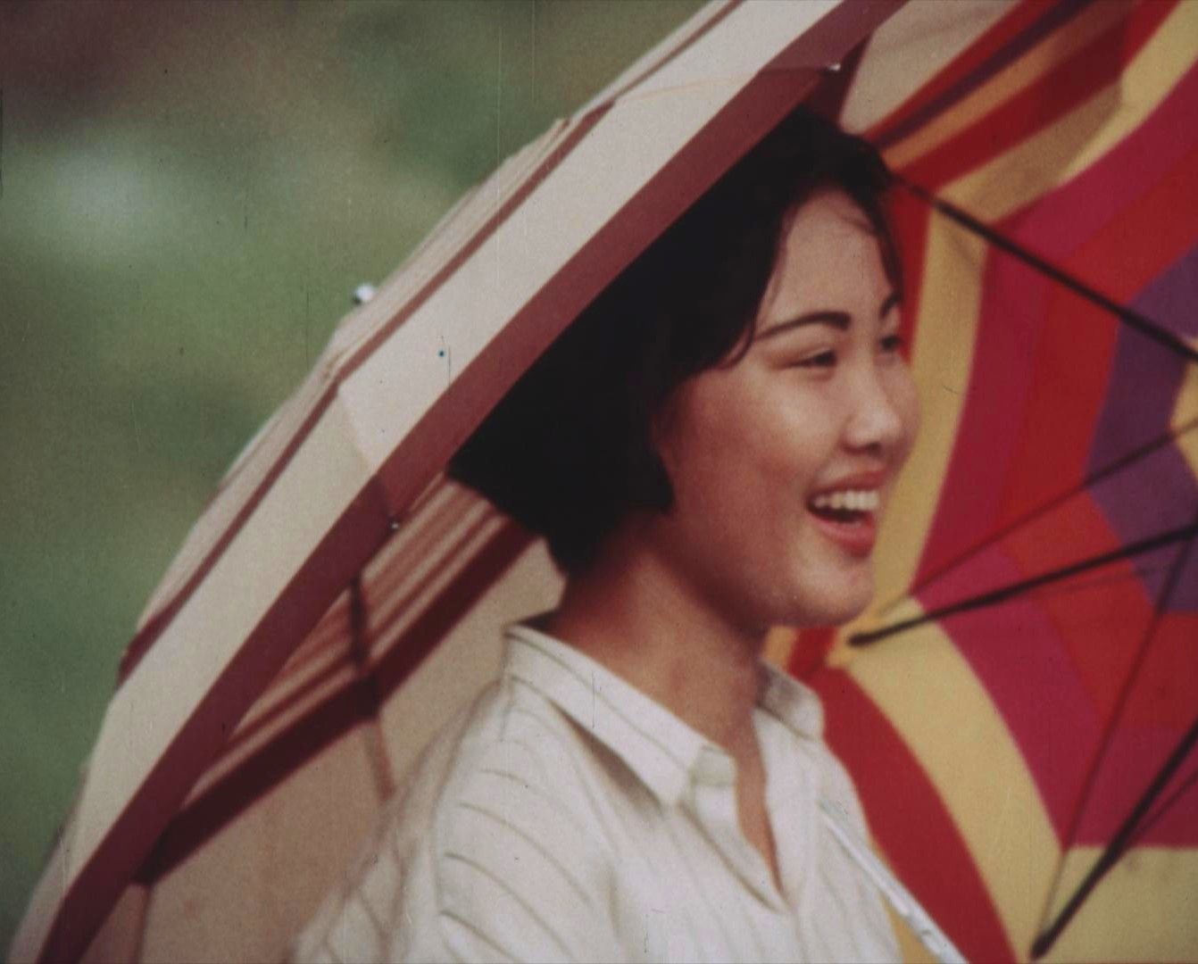 A young Vietnamese woman wearing a white blouse with black stripes. She is smiling and carrying an open umbrella with brown stripes. Next to her is an open umbrella with red, yellow and purple stripes.