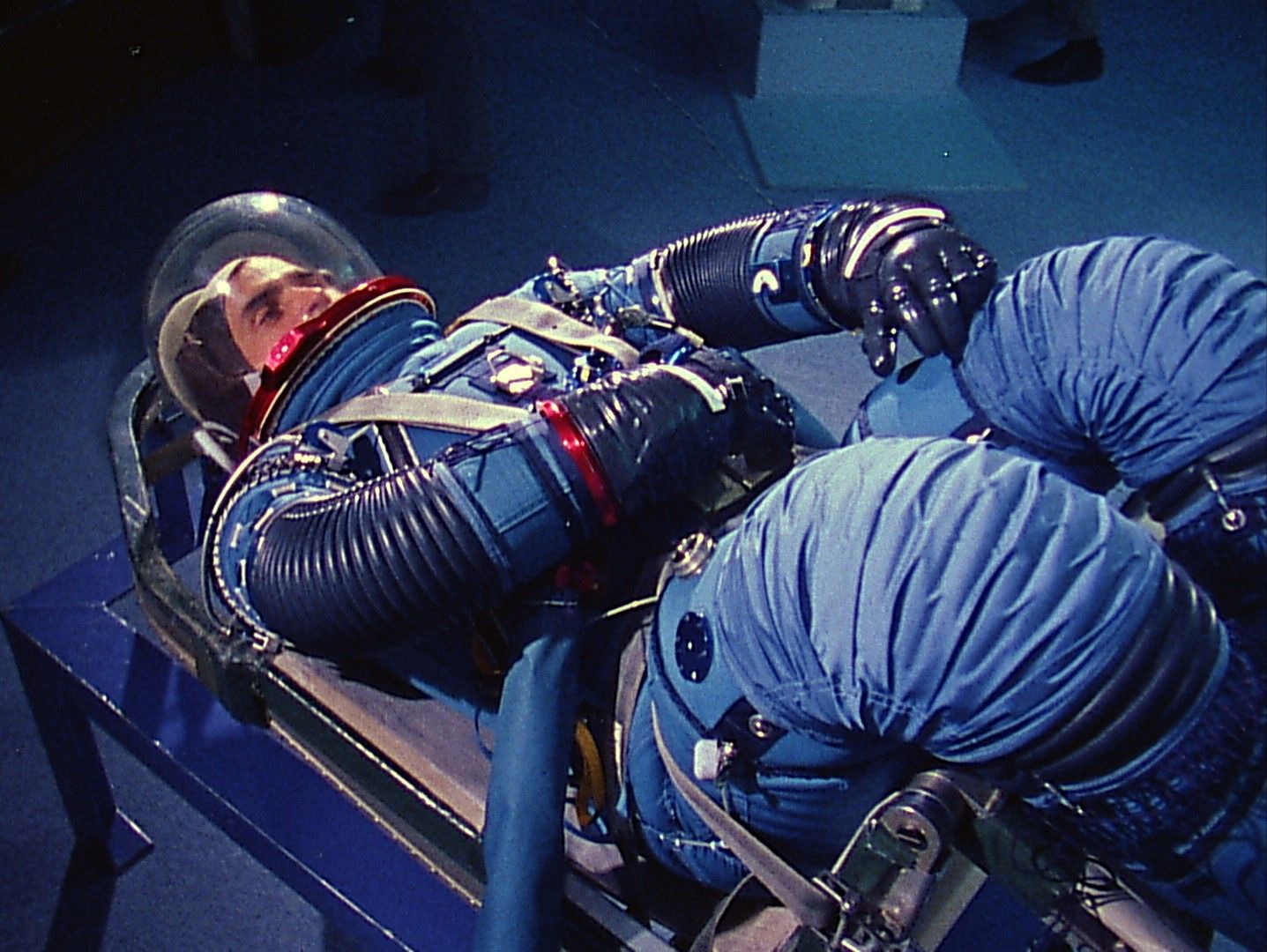 Astronaut Harrison Schmitt lies horizontal, strapped to a seat in a NASA test facility. He is wearing a blue space suit and helmet.