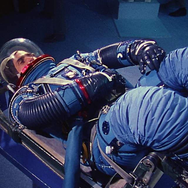 Outer Space | Space Race / Weltraum | Wettlauf ins All - Apollo 17: Ein Sonderbericht (H00221) Astronaut Harrison Schmitt lies horizontal, strapped to a seat in a NASA test facility. He is wearing a blue space suit and helmet.