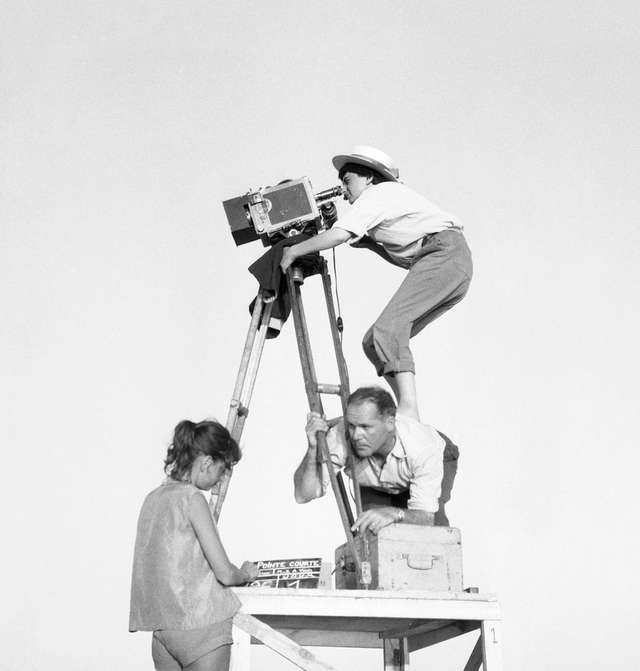 Director Agnès Varda stands on the back of her cameraman and looks through the camera.