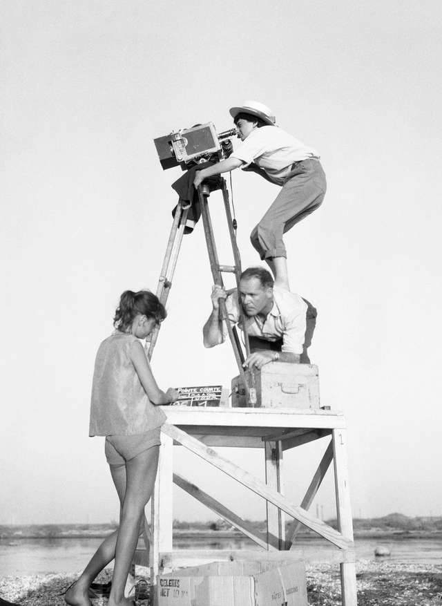 Director Agnès Varda stands on the back of her cameraman and looks through the camera.
