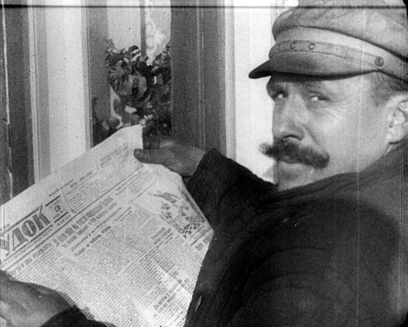 Black-and-white image of an older man with a moustache and a flat cap. He is holding an open newspaper in his hands and looking directly into the camera.