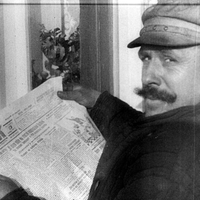 VUFKU-Filmwoche 1927/44 (UA00006) Black-and-white image of an older man with a moustache and a flat cap. He is holding an open newspaper in his hands and looking directly into the camera.