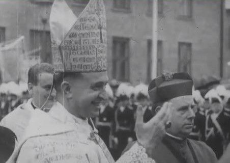 Black and white photograph: close-up of Archbishop Aloysius Muench in clerical dress and pointed bishop's mitre, inscribed with medieval script. Standing next to him is a cardinal wearing a cardinal's cap.