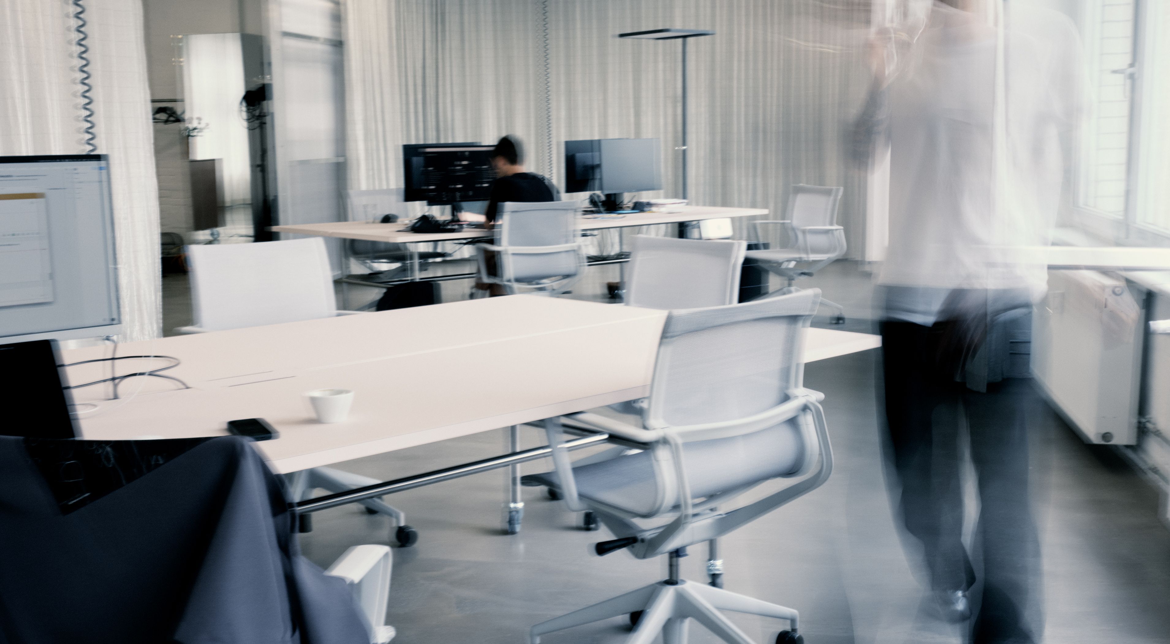 Blurred figure walking through a modern open-plan office with white desks, rolling chairs, and computer monitors, while another person works at a desk.