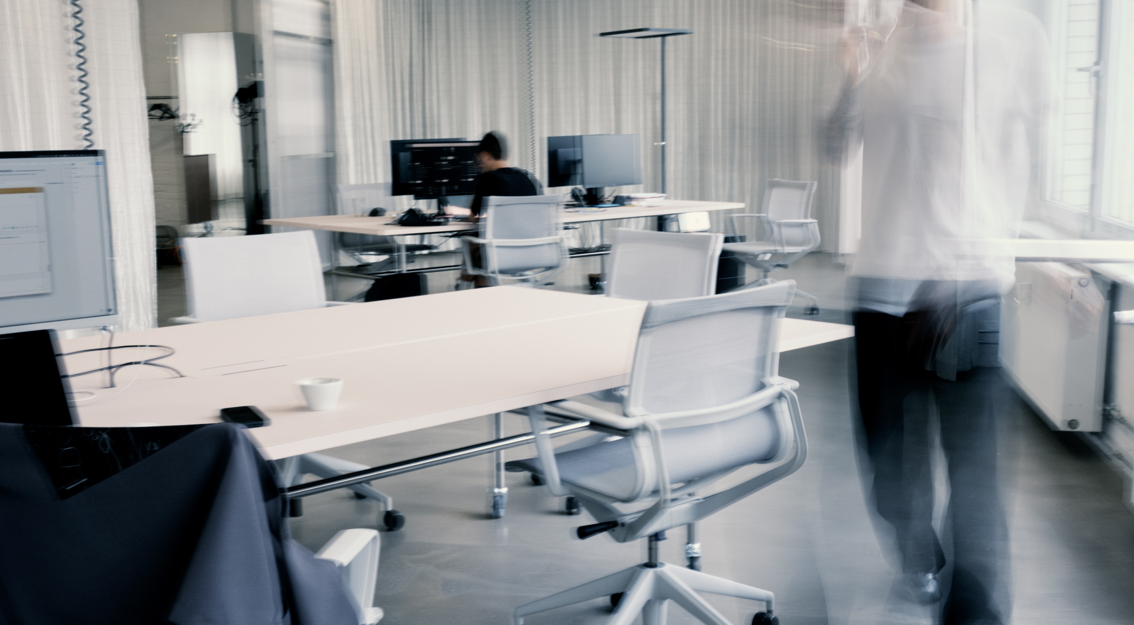 Blurred figure walking through a modern open-plan office with white desks, rolling chairs, and computer monitors, while another person works at a desk.