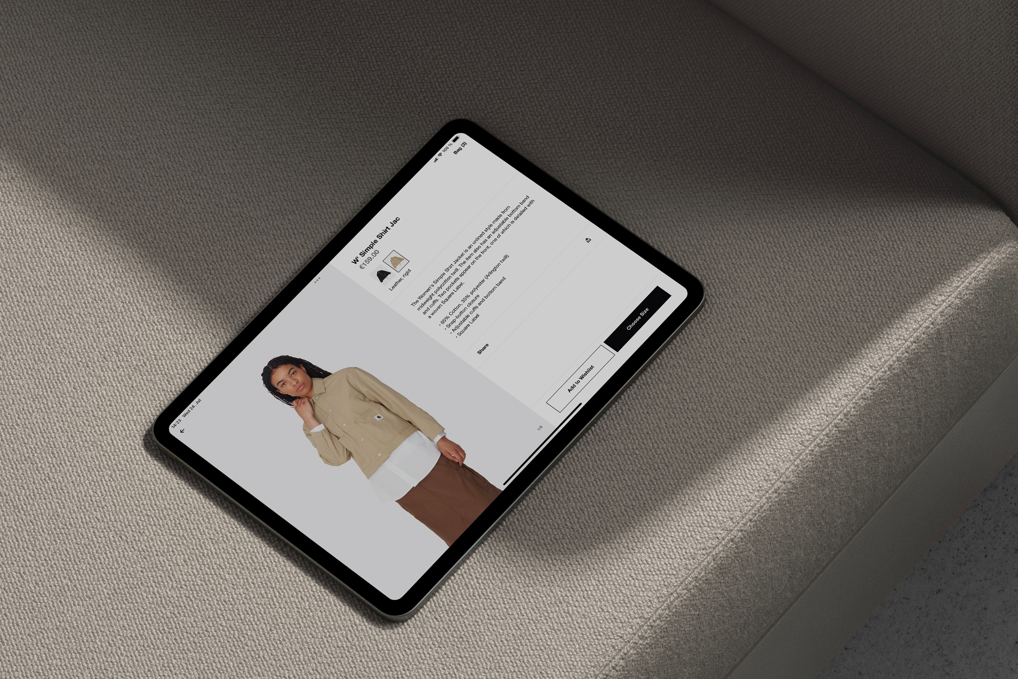 Tablet on a textured gray sofa displaying an online clothing store page with a model wearing a beige jacket.