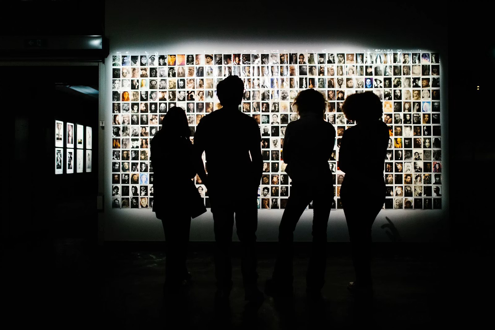 Silhouettes of four people stand in front of a brightly lit wall covered with many small portrait photographs in a dark gallery.
