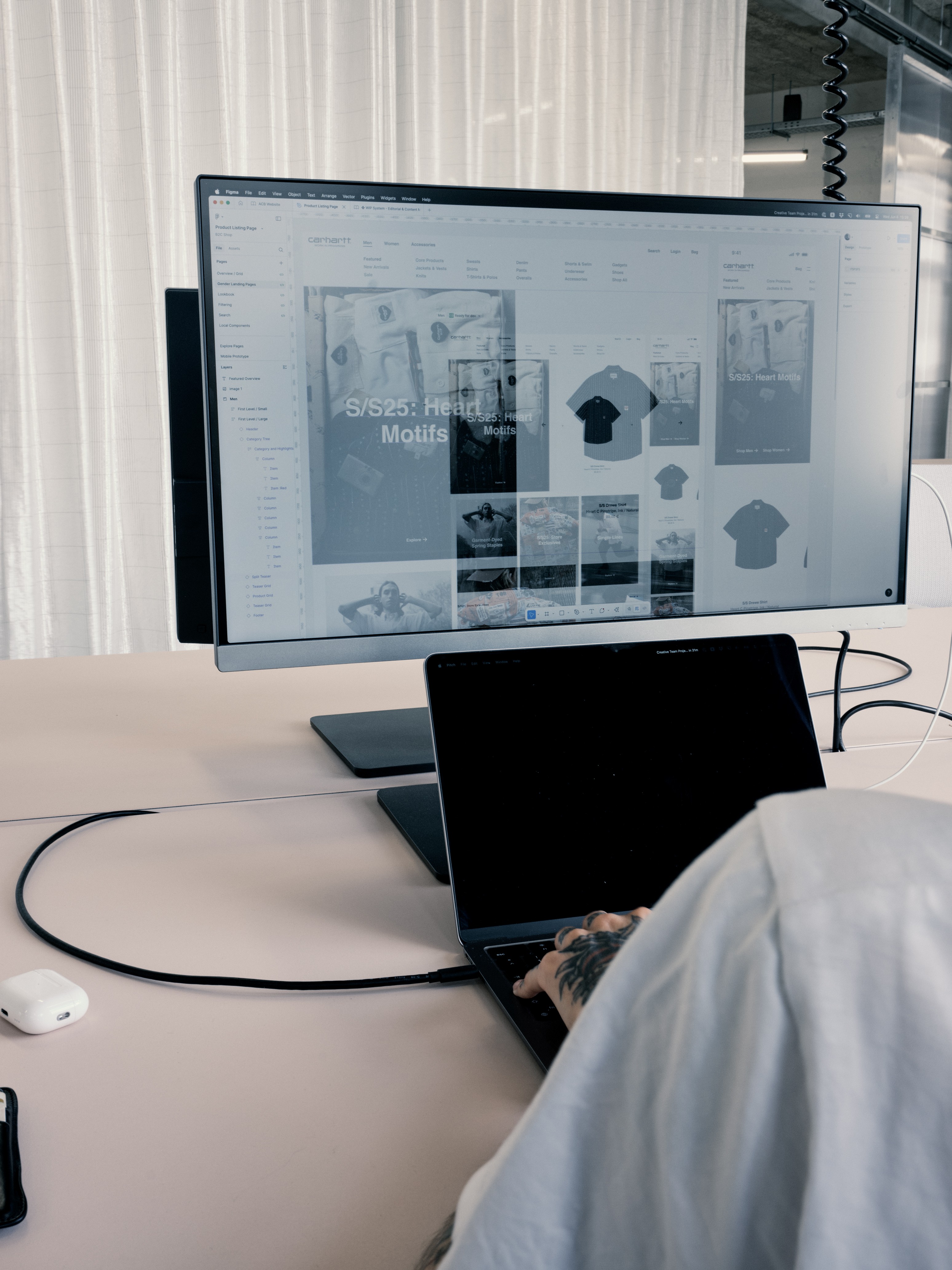 Person working on a laptop connected to a large monitor displaying a clothing website design in a modern office.