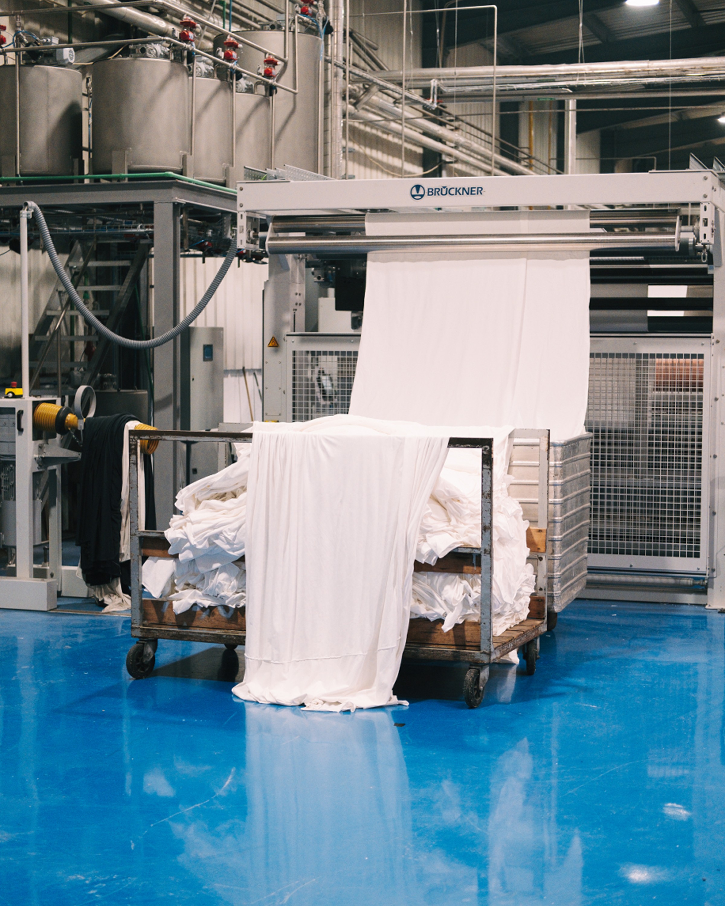 Industrial textile machine feeding white fabric into a cart piled with cloth on a shiny blue factory floor.