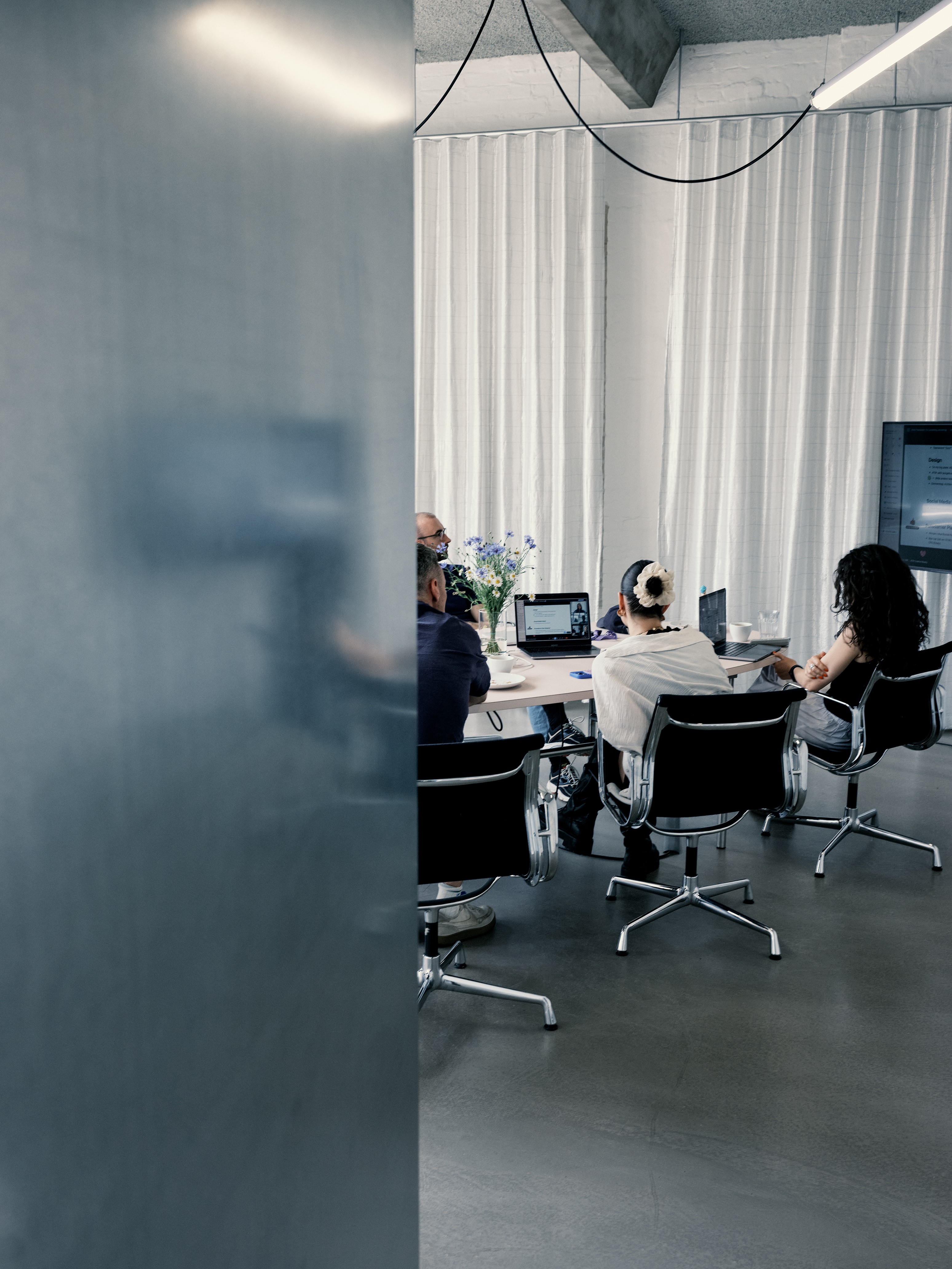 People sit around a conference table with laptops in a modern office, viewed partially from behind a wall.