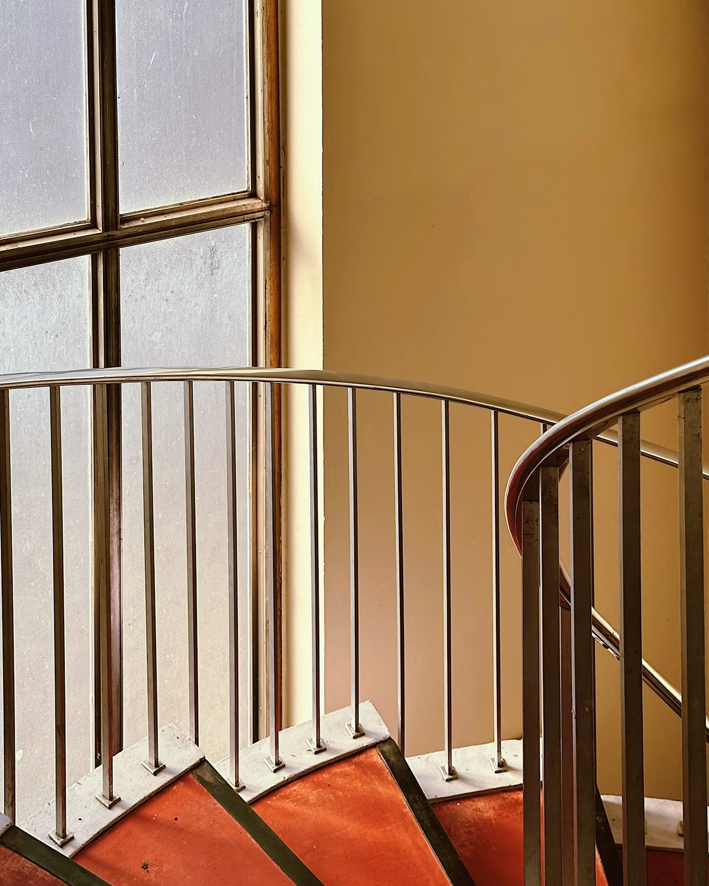 Curved indoor staircase with metal railings, red steps, warm beige walls, and tall frosted windows.