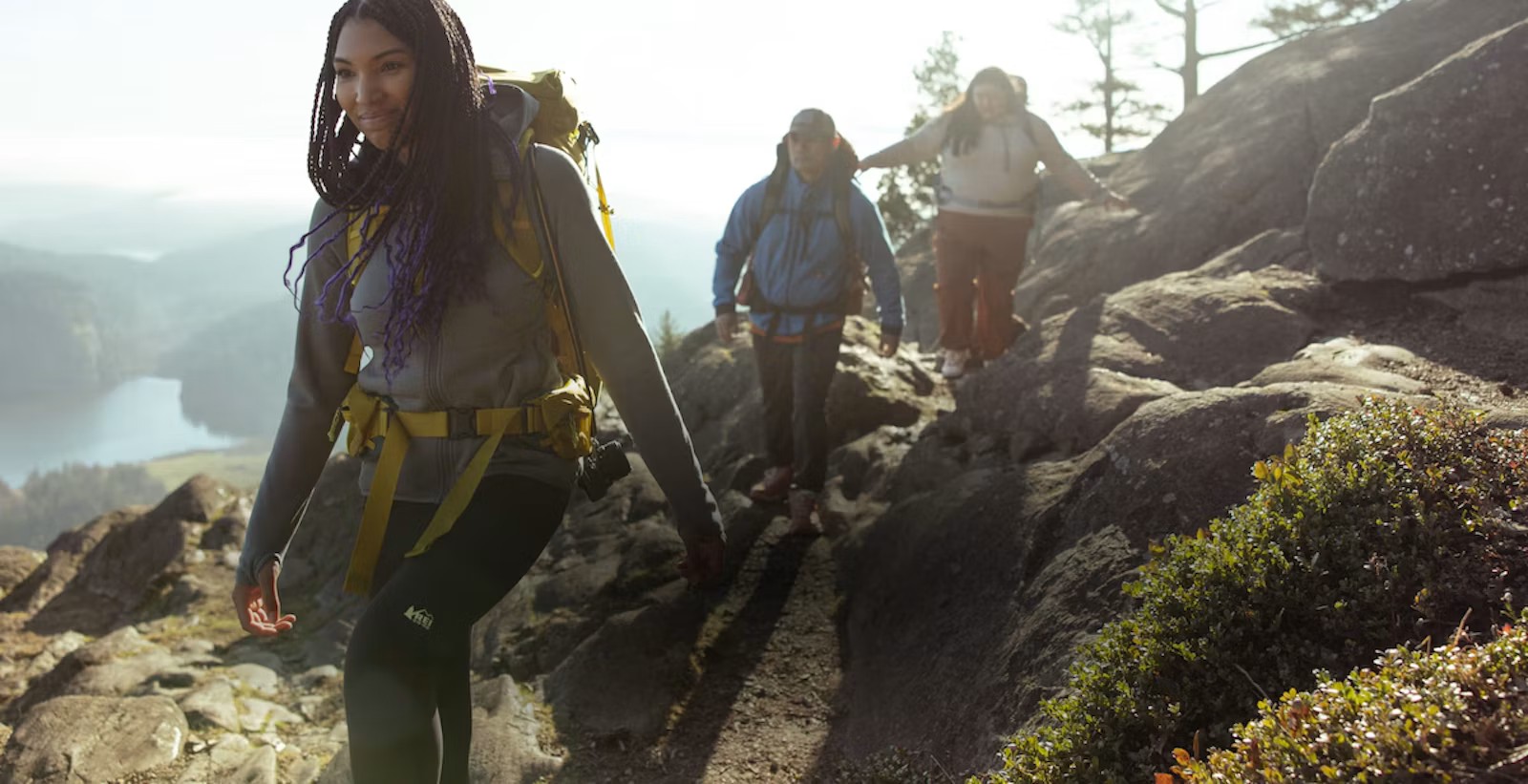 Three hikers with backpacks walk along a rocky mountain trail above a lake on a sunny day.