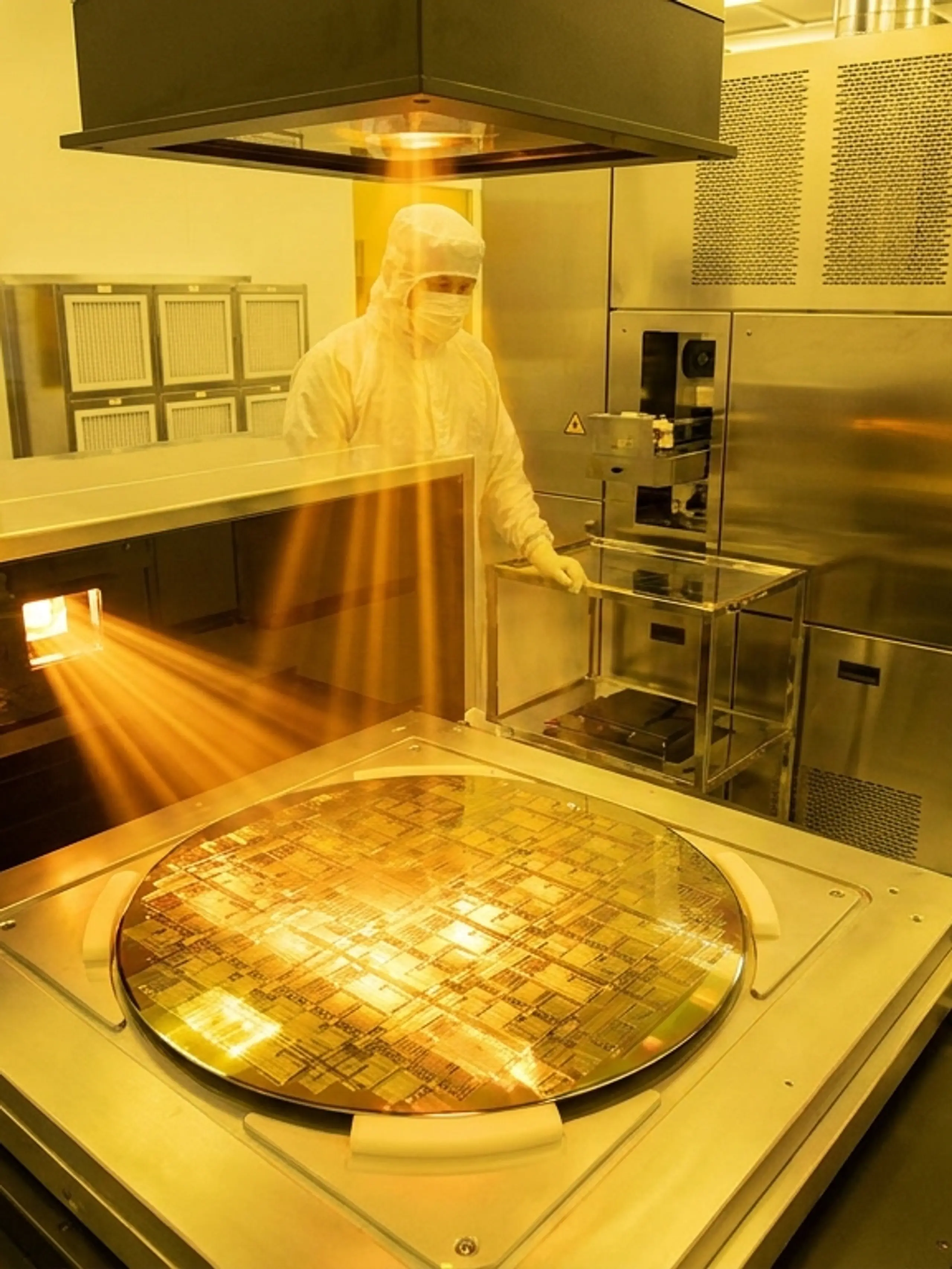 A silicon wafer being processed by technicians in a semiconductor cleanroom under amber light.