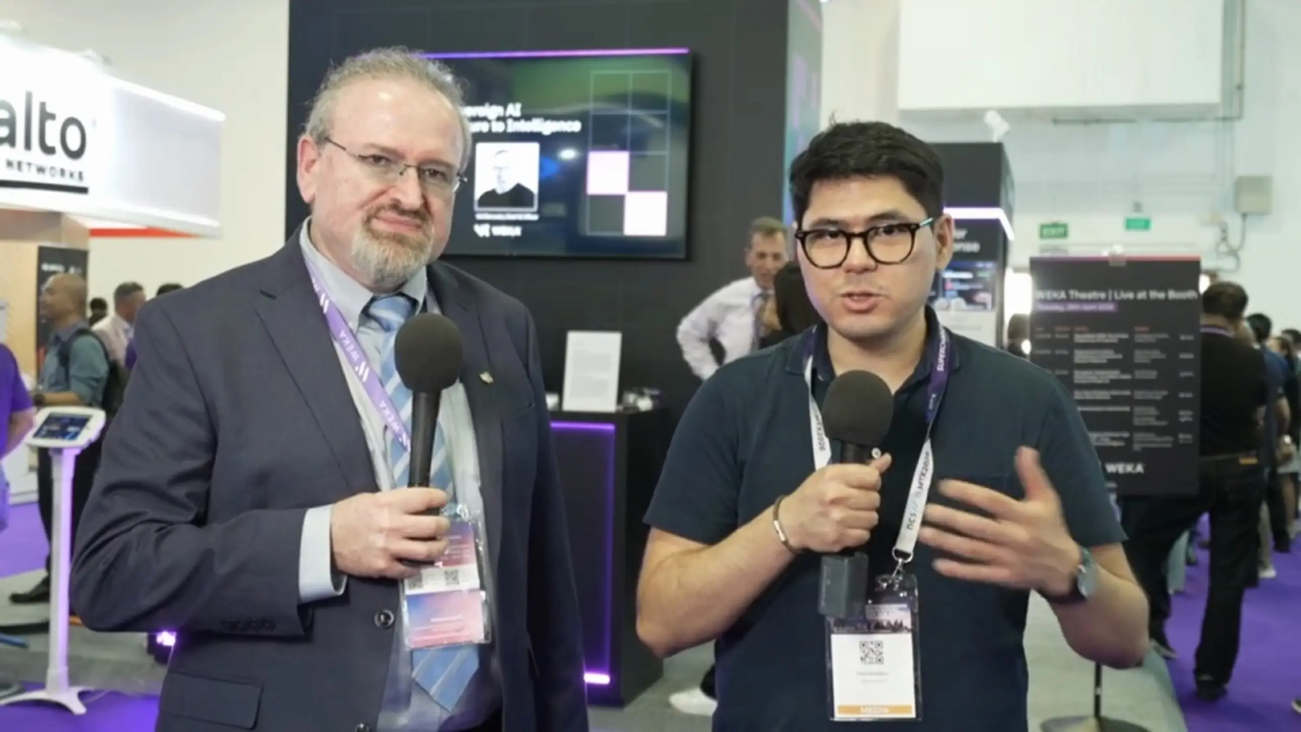 Two men holding microphones, one older in a suit and one younger in a polo shirt, present at a technology exhibition booth.
