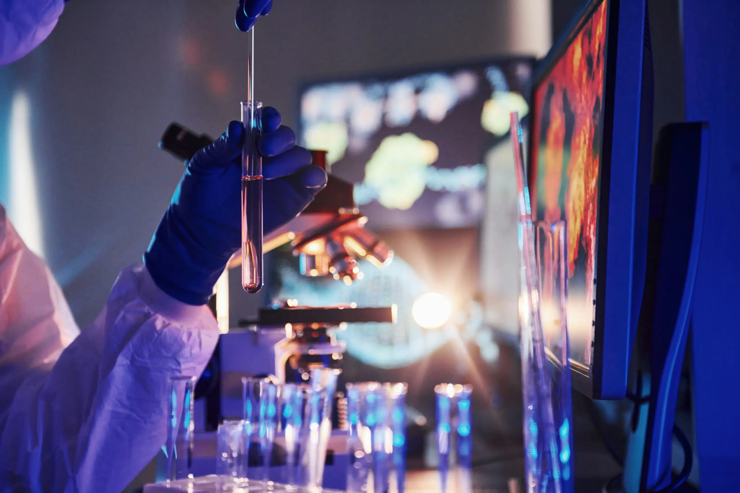 A gloved hand holds a test tube in a blue-lit science lab with a microscope and digital screens.