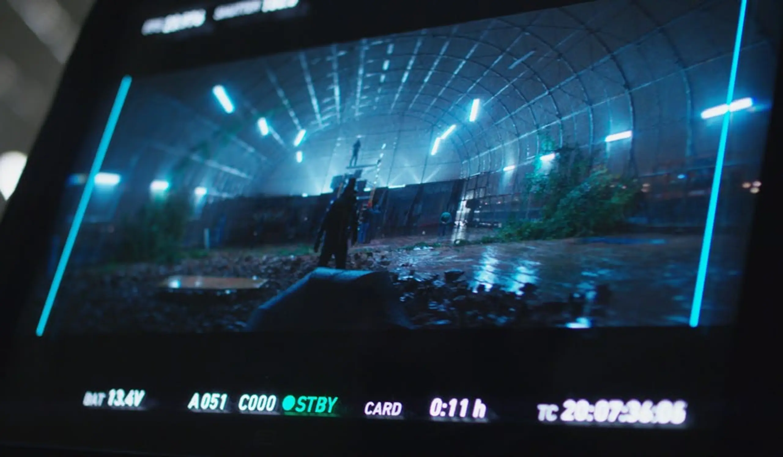 Camera monitor displaying a dark, wet, futuristic tunnel scene with blue lighting and two figures.