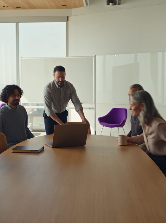 Four people in a modern meeting room, with one man standing by a laptop and others seated at a large table.