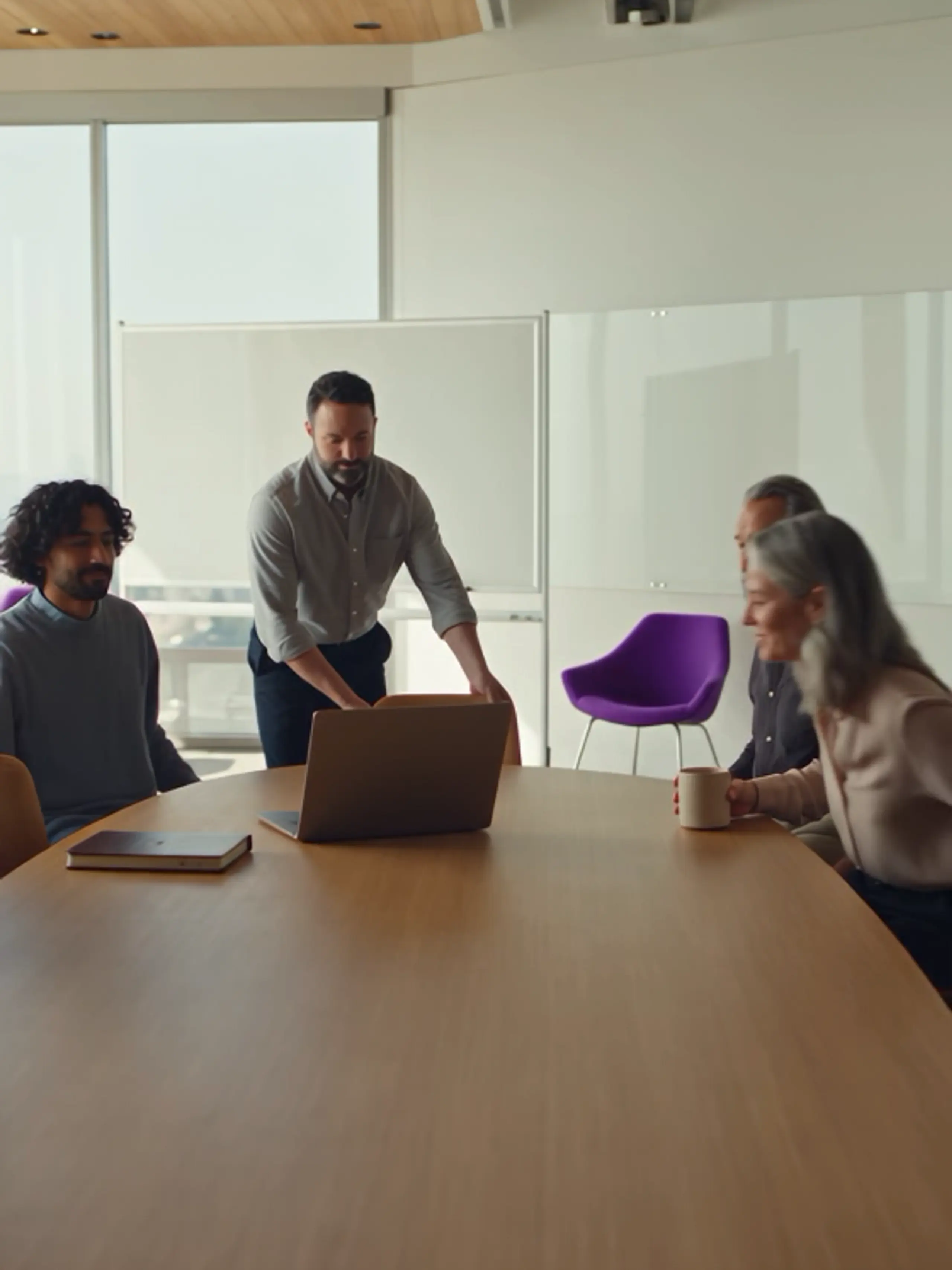 Four people in a modern meeting room, with one man standing by a laptop and others seated at a large table.