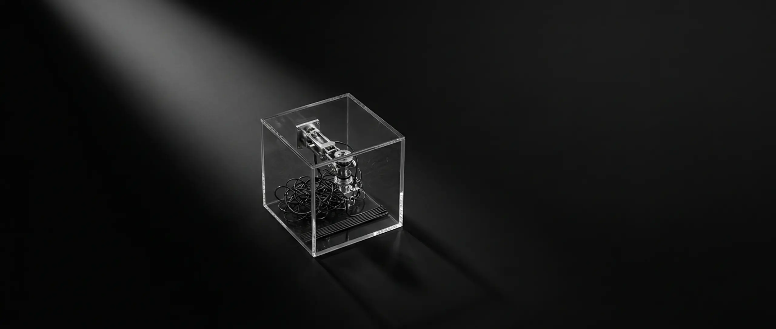 A clear acrylic cube holds an intricate metallic mechanism and black coils, spotlighted on a dark surface.