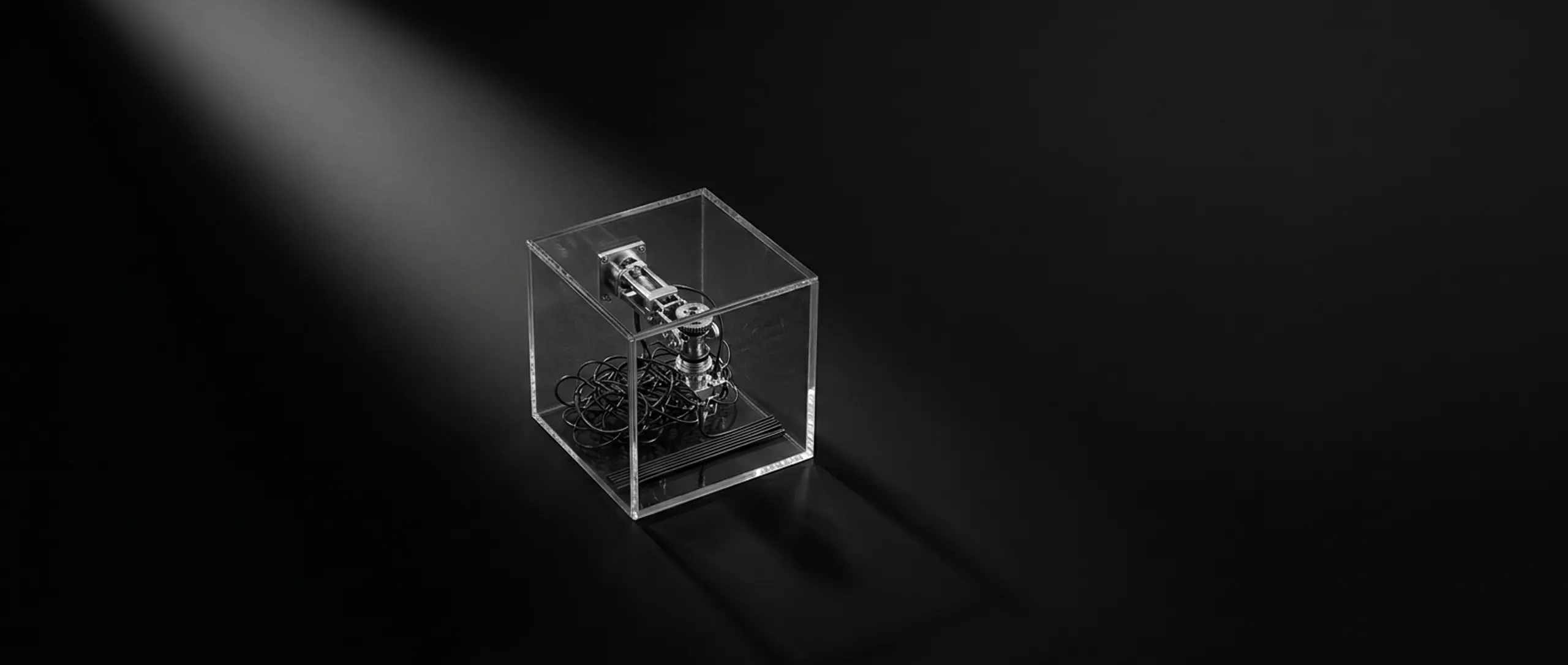 A clear acrylic cube holds an intricate metallic mechanism and black coils, spotlighted on a dark surface.
