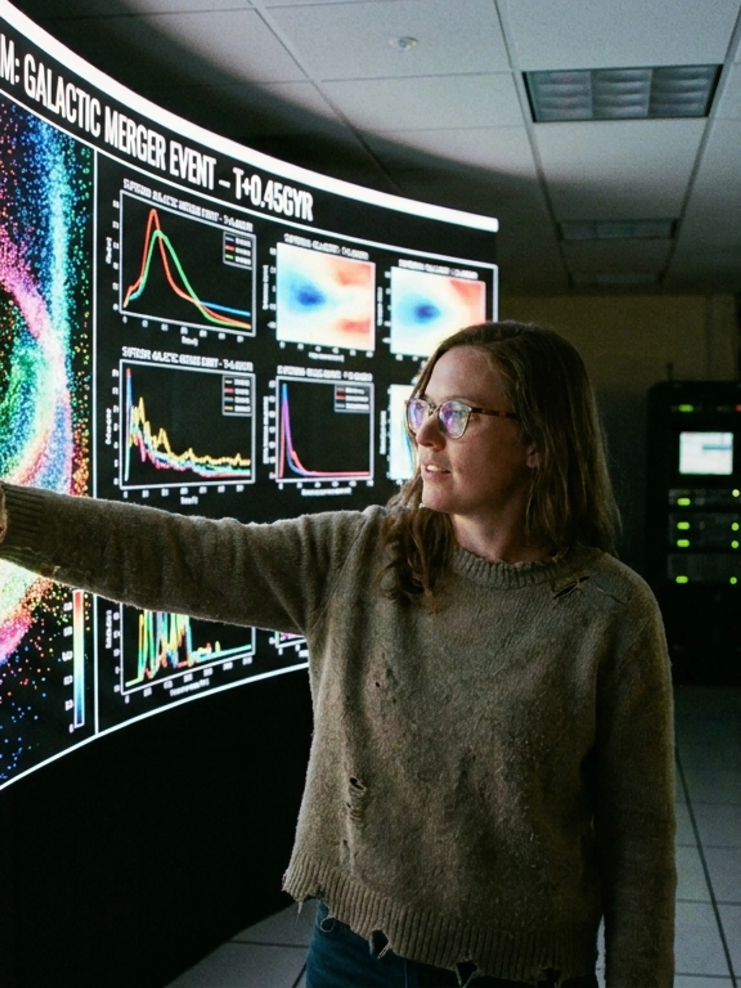 A woman points at a large curved screen displaying a colorful galactic merger simulation and data graphs in a server room.
