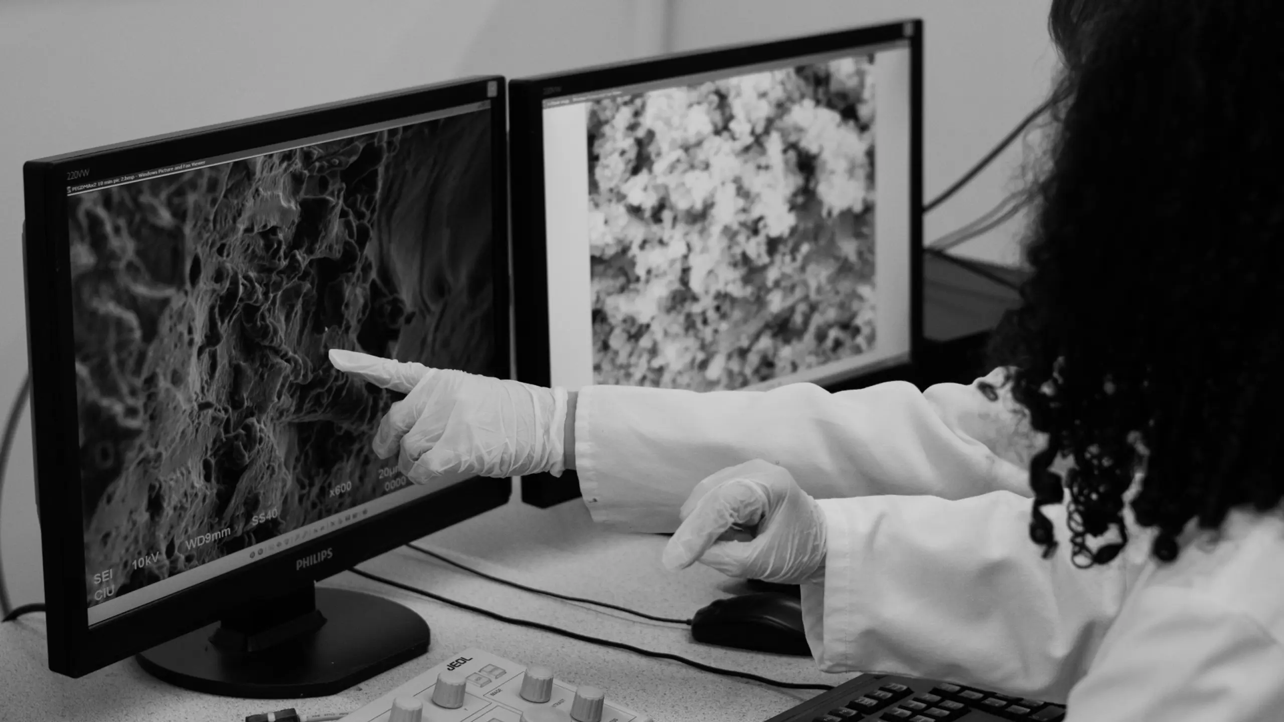 A person in a lab coat and gloves points at a monitor displaying a magnified, textured black and white scientific image.