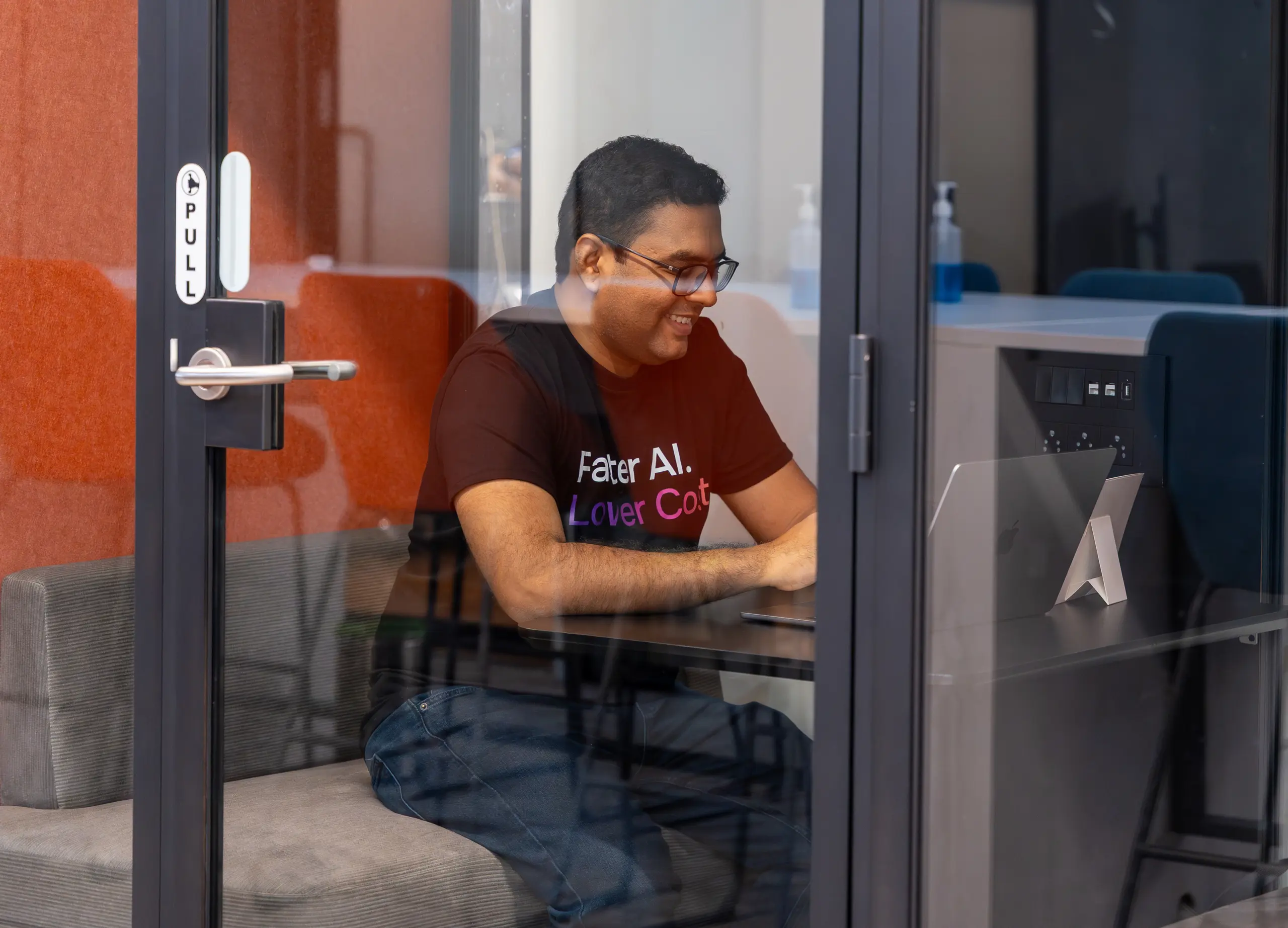 A smiling man in glasses works on a laptop in a glass-walled office pod.