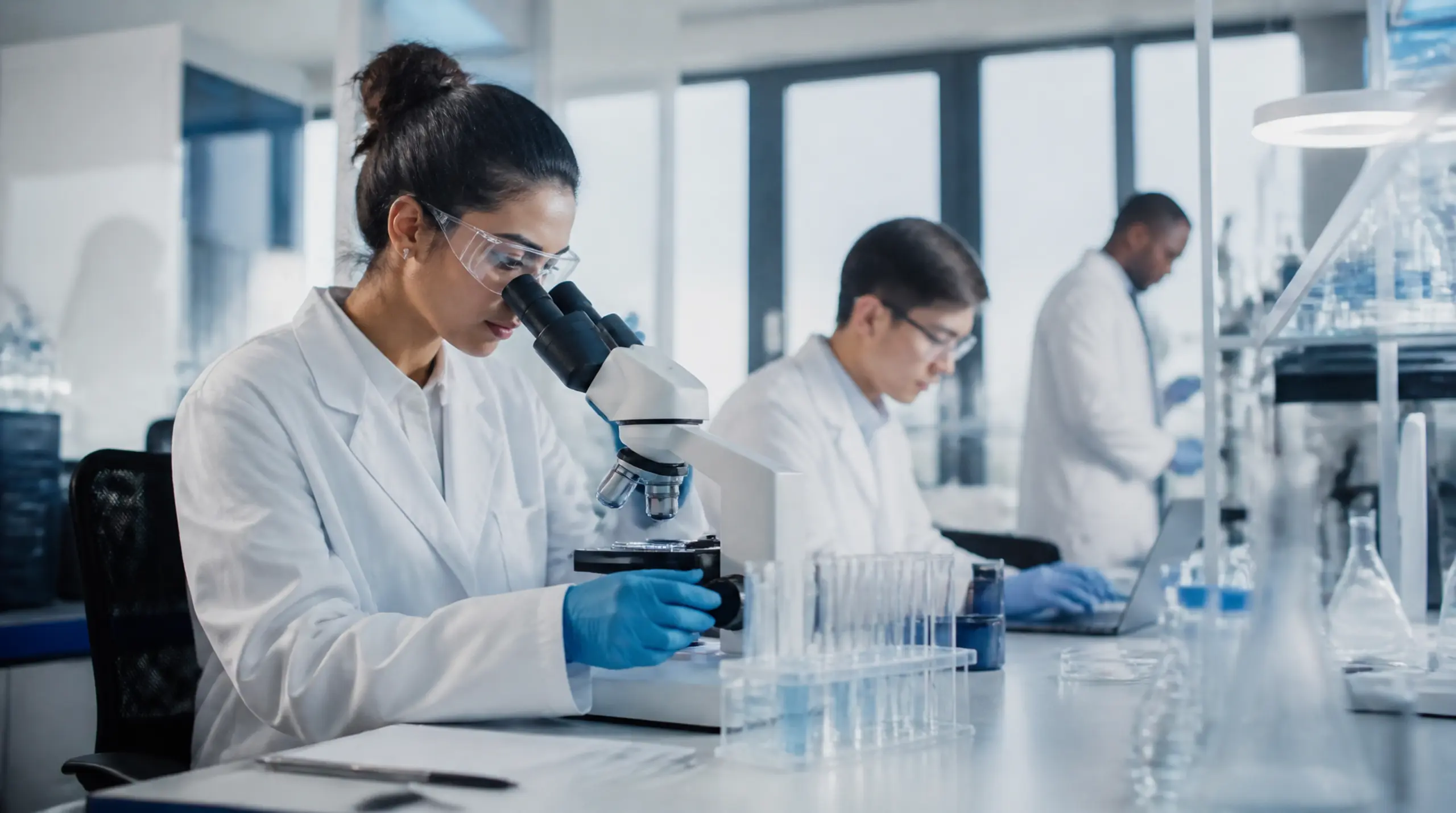 A scientist looks through a microscope in a laboratory while colleagues work nearby.