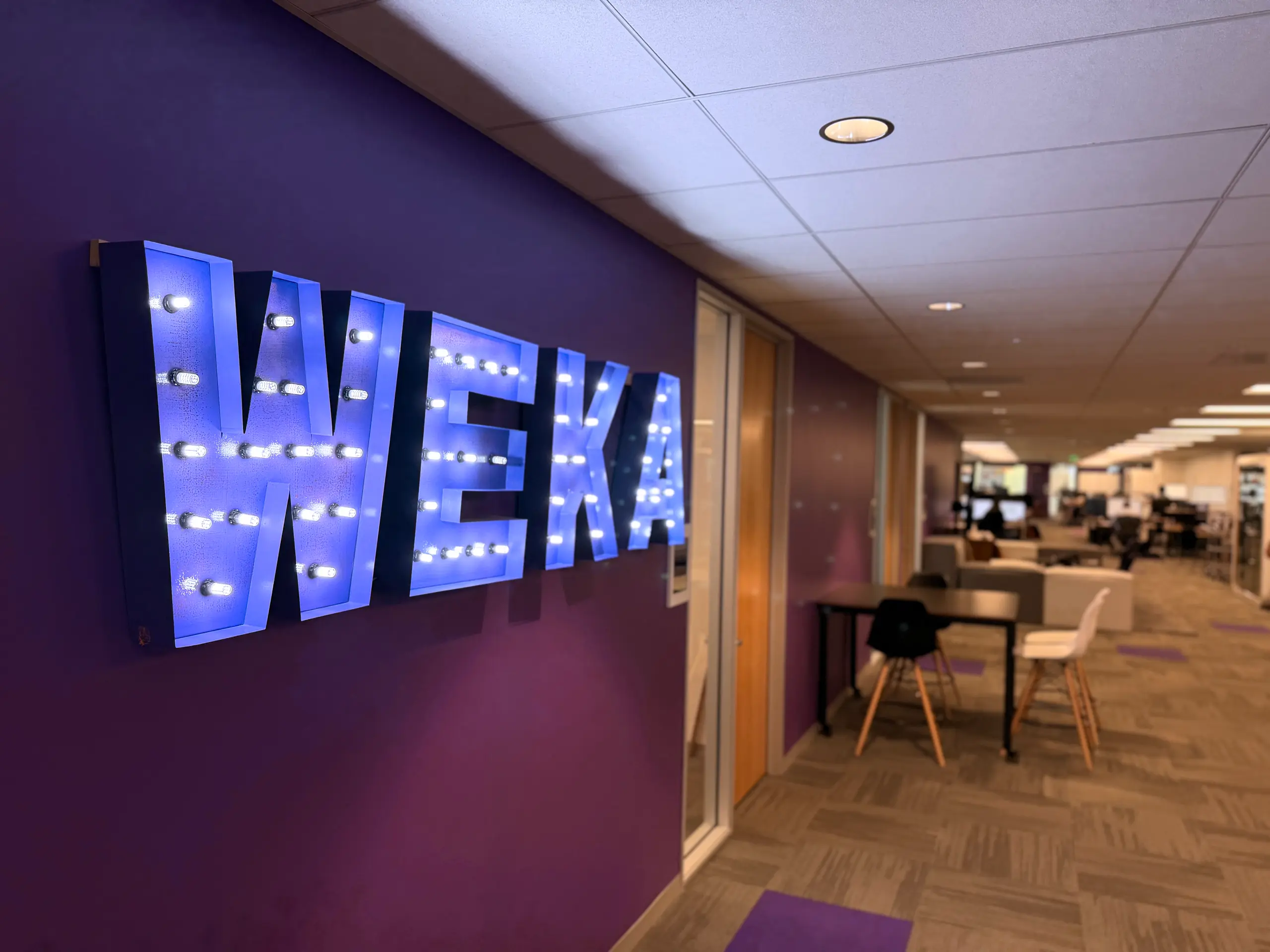 A large, purple, illuminated "WEKA" sign on a purple wall in a modern office hallway.