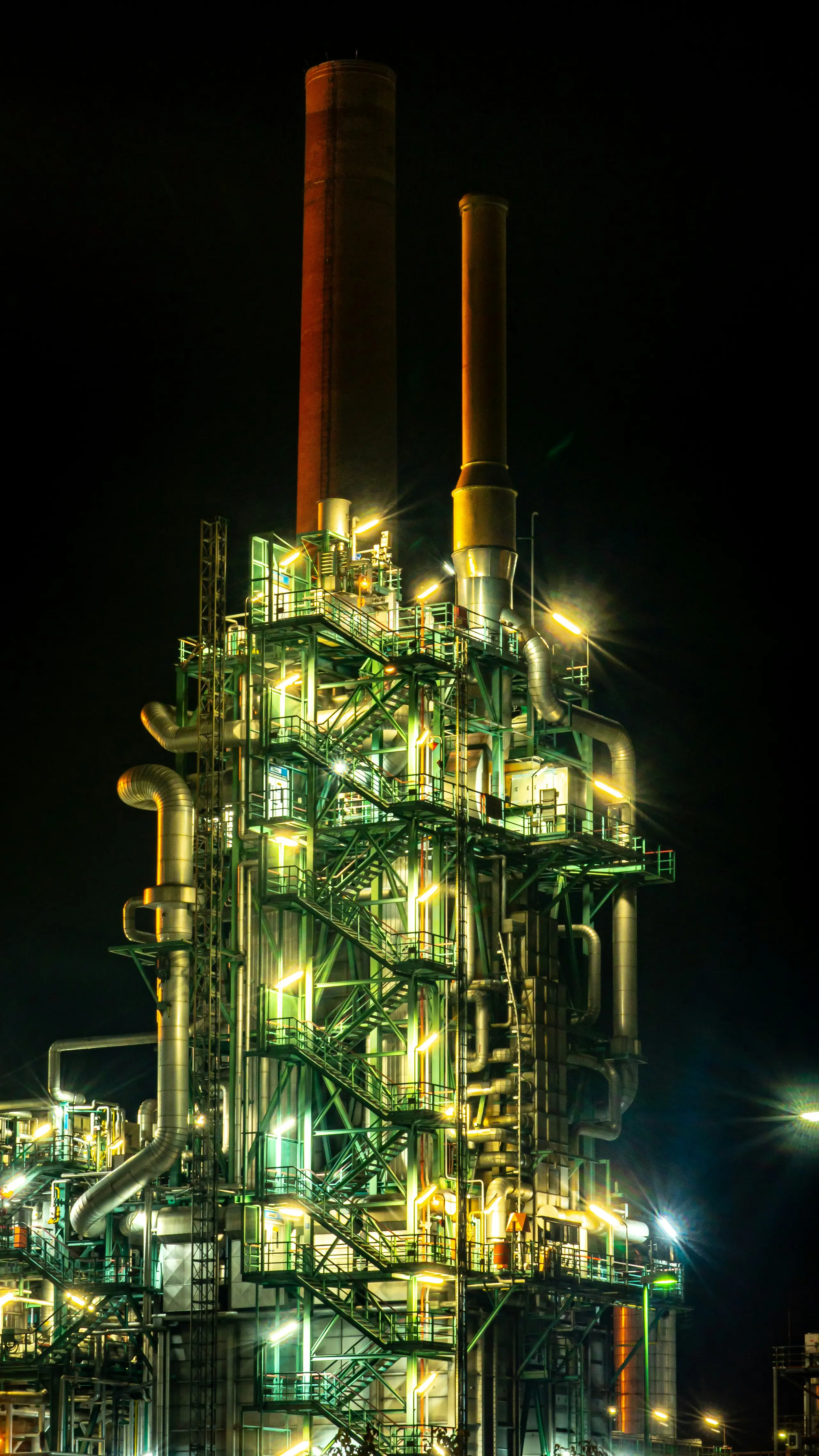 A brightly lit industrial plant at night, with a complex structure of pipes and platforms, and two tall smokestacks against a dark sky.