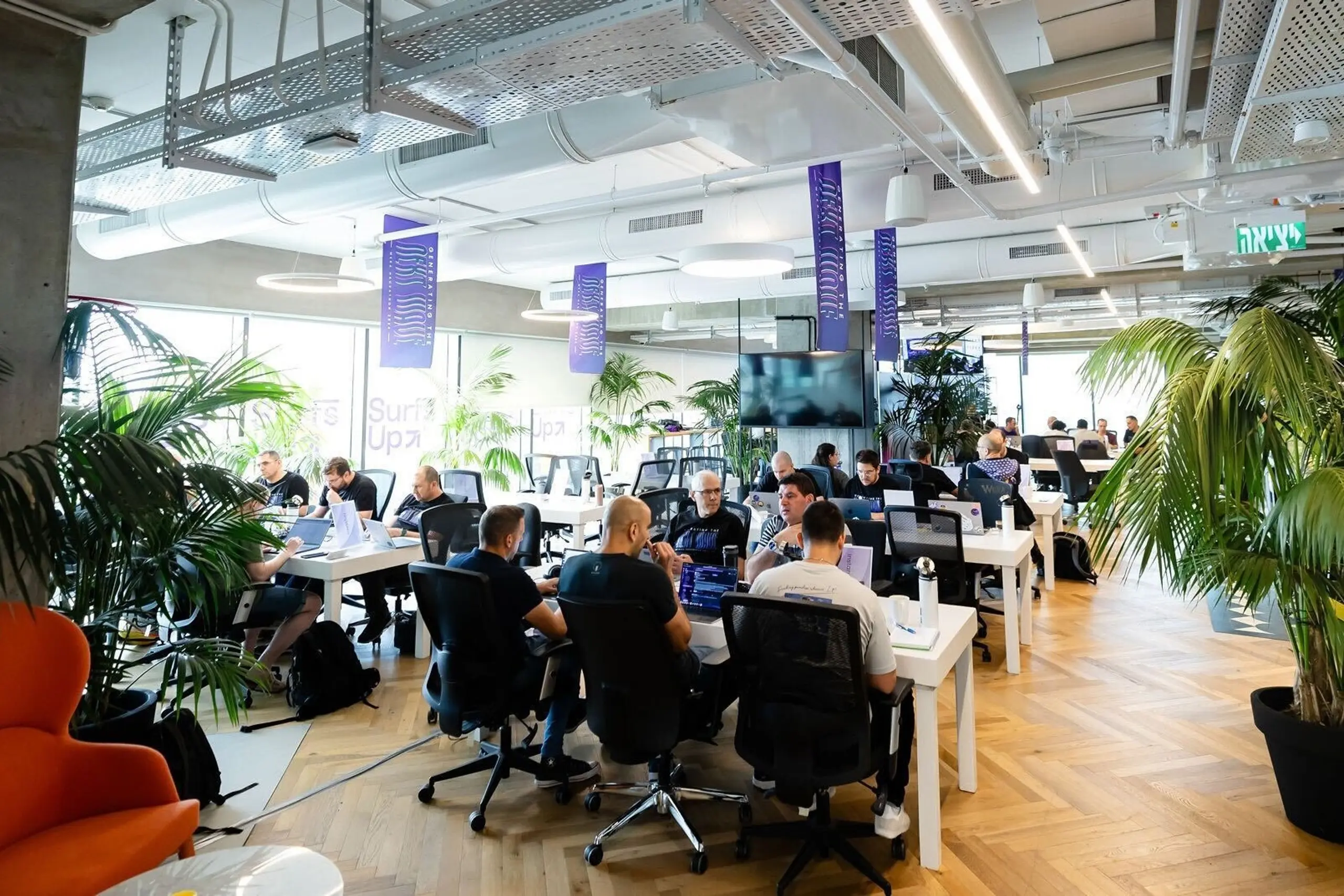 A modern, open-plan office with people working at desks, potted plants, and an exposed ceiling.