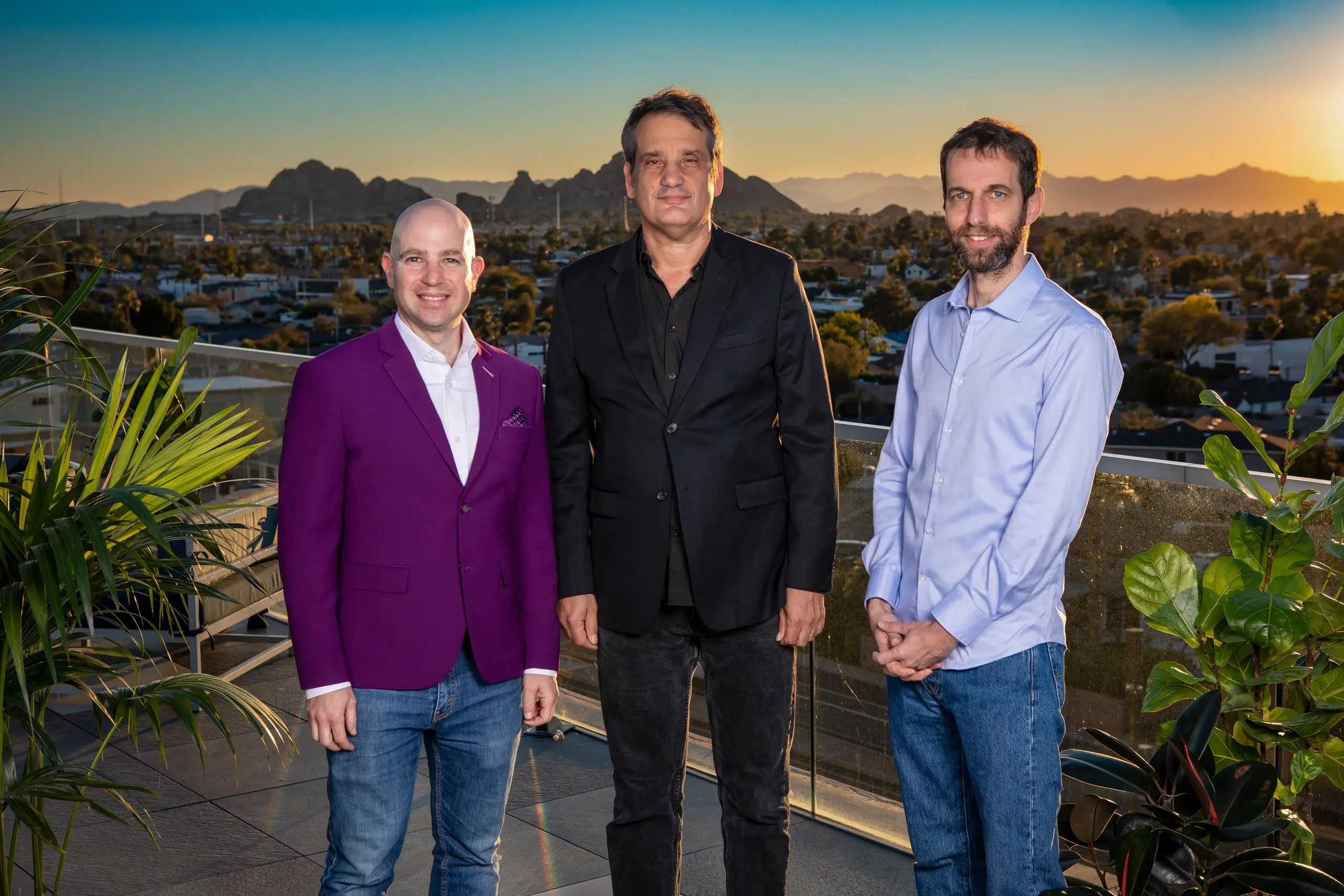 Three men stand on a rooftop overlooking a city and mountains at sunset.