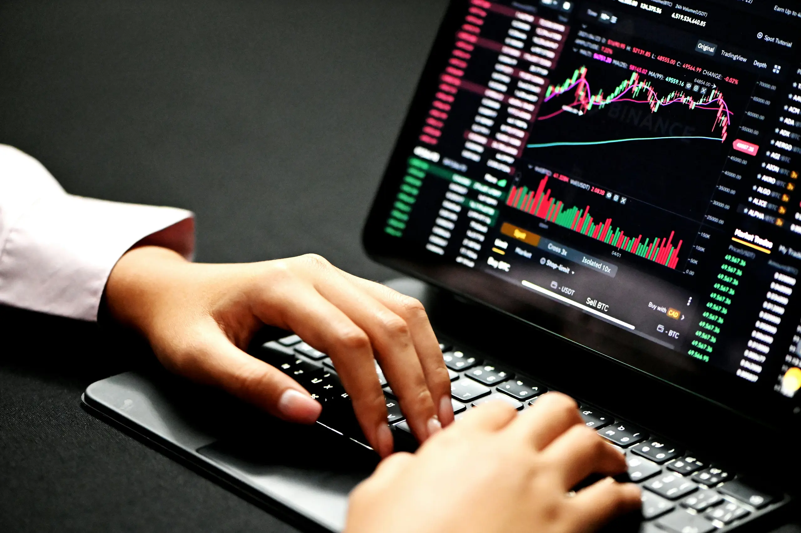 Hands typing on a laptop displaying cryptocurrency trading charts.