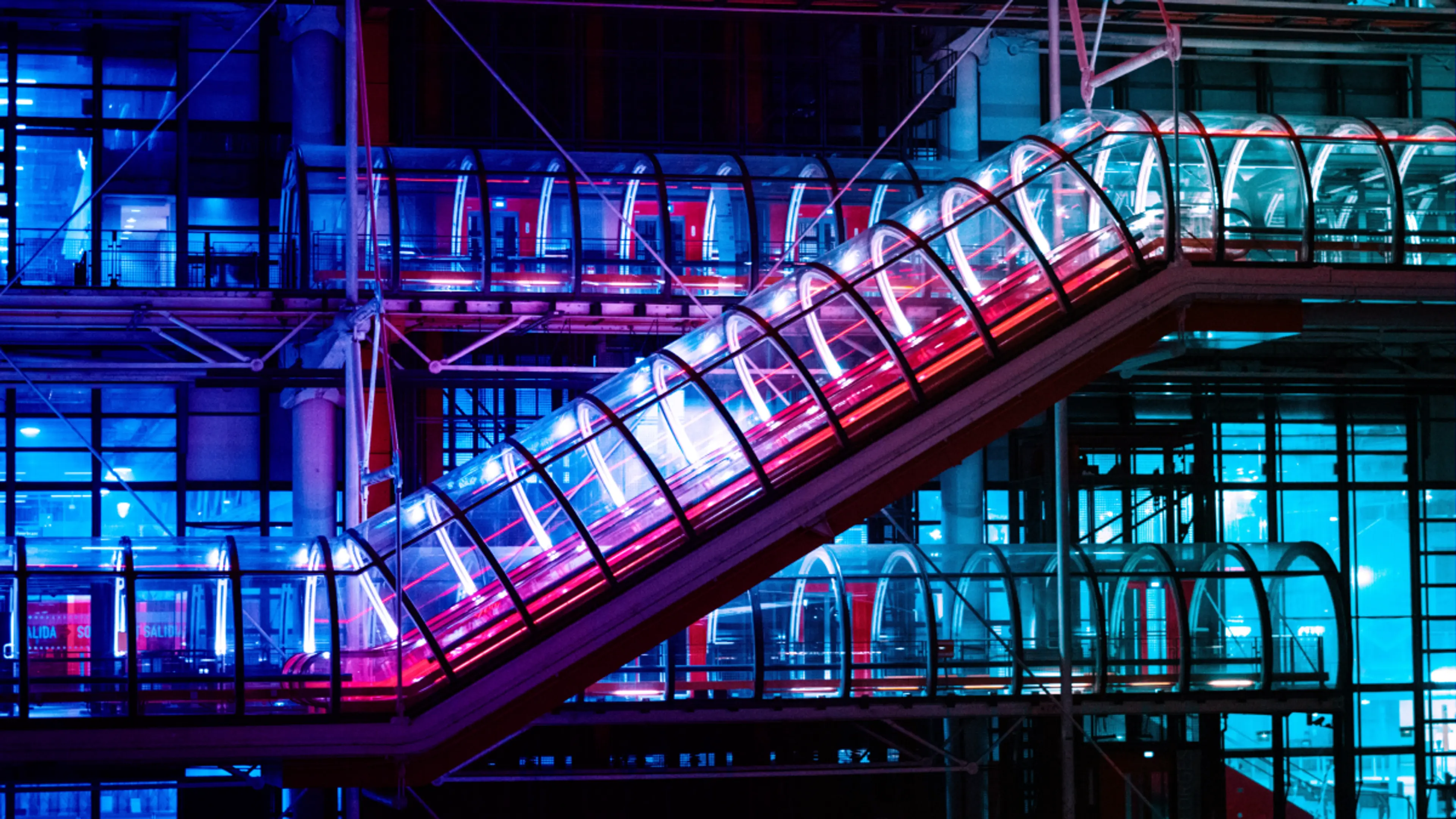 External glass escalator and walkways on a modern building, illuminated in vibrant blue and pink light.
