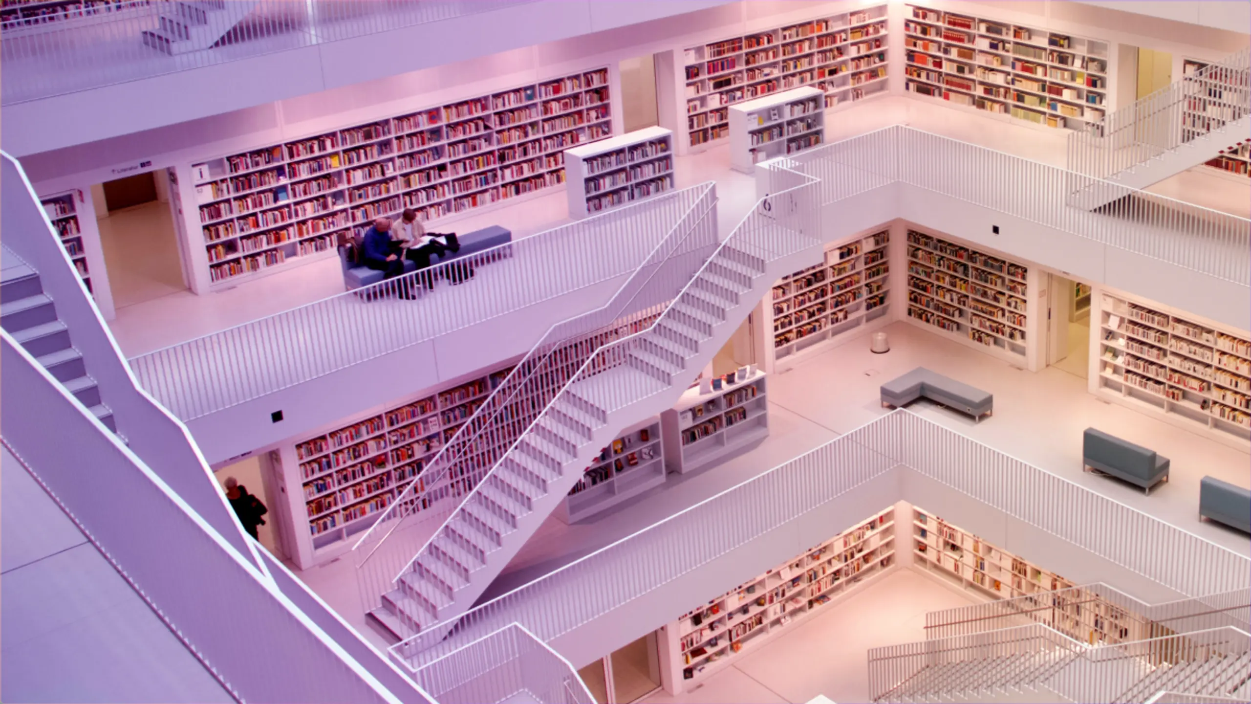 Multi-level white library interior with bookshelves, intersecting staircases, and a few people.