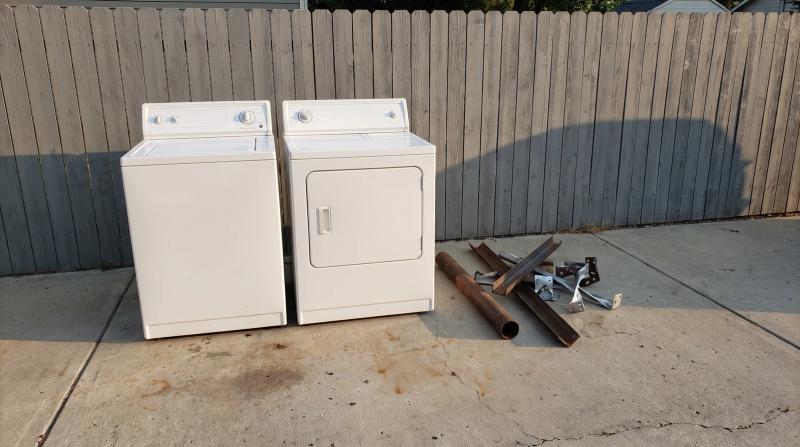 An old washer and dryer set on a concrete driveway beside a stack of scrap metal, ready for appliance removal at a home in Nampa, Idaho.