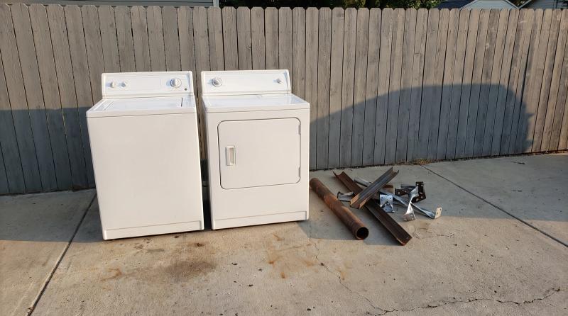 An old washer and dryer set on a concrete driveway beside a stack of scrap metal, ready for appliance removal at a home in Nampa, Idaho.