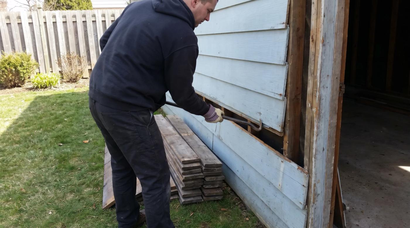 A worker in plain clothes prying a weathered board off the frame of a small old detached garage in Boise, Idaho, with salvaged lumber stacked on the grass nearby.