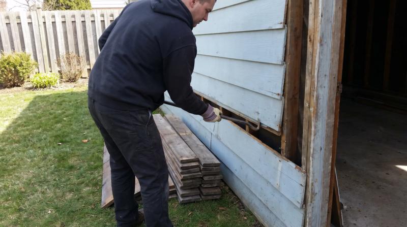 A worker in plain clothes prying a weathered board off the frame of a small old detached garage in Boise, Idaho, with salvaged lumber stacked on the grass nearby.