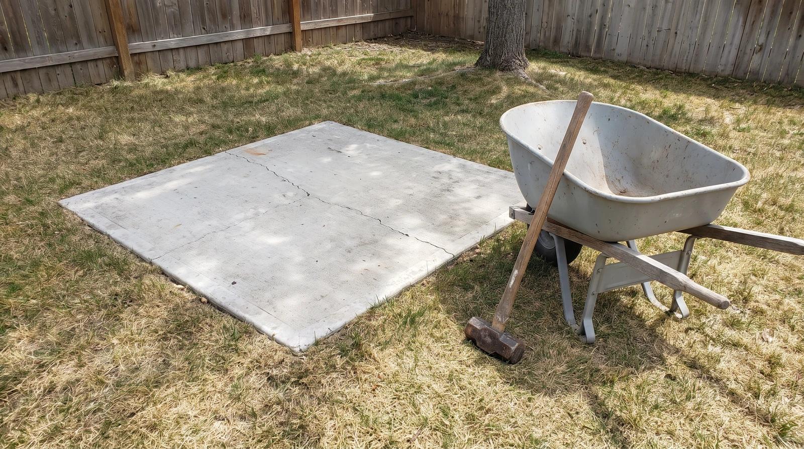 Empty cracked concrete hot tub pad in an Emmett, Idaho backyard with a sledgehammer and wheelbarrow beside it, ready to be broken out after hot tub removal