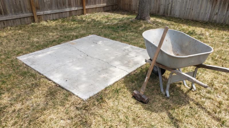 Empty cracked concrete hot tub pad in an Emmett, Idaho backyard with a sledgehammer and wheelbarrow beside it, ready to be broken out after hot tub removal
