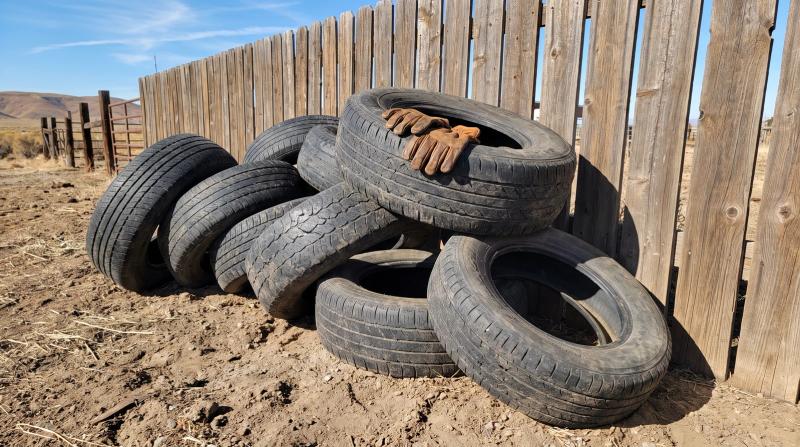 Old weathered tire pile half buried in dirt beside a wooden ranch fence, waiting for bulk tire disposal on a rural property near Kuna, Idaho
