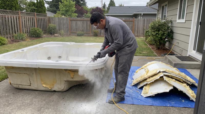 Worker cutting apart an old fiberglass hot tub shell with a reciprocating saw on a backyard patio in Garden City, Idaho