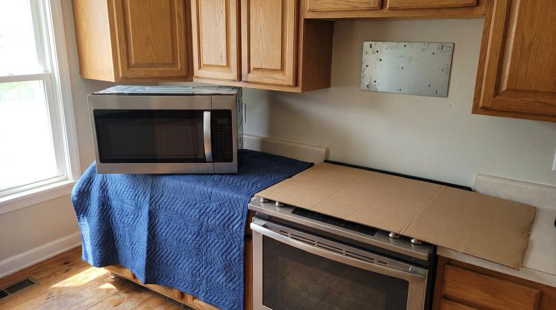 Old over-the-range microwave on a moving blanket on a kitchen floor below an empty wall mounting plate and a covered stovetop