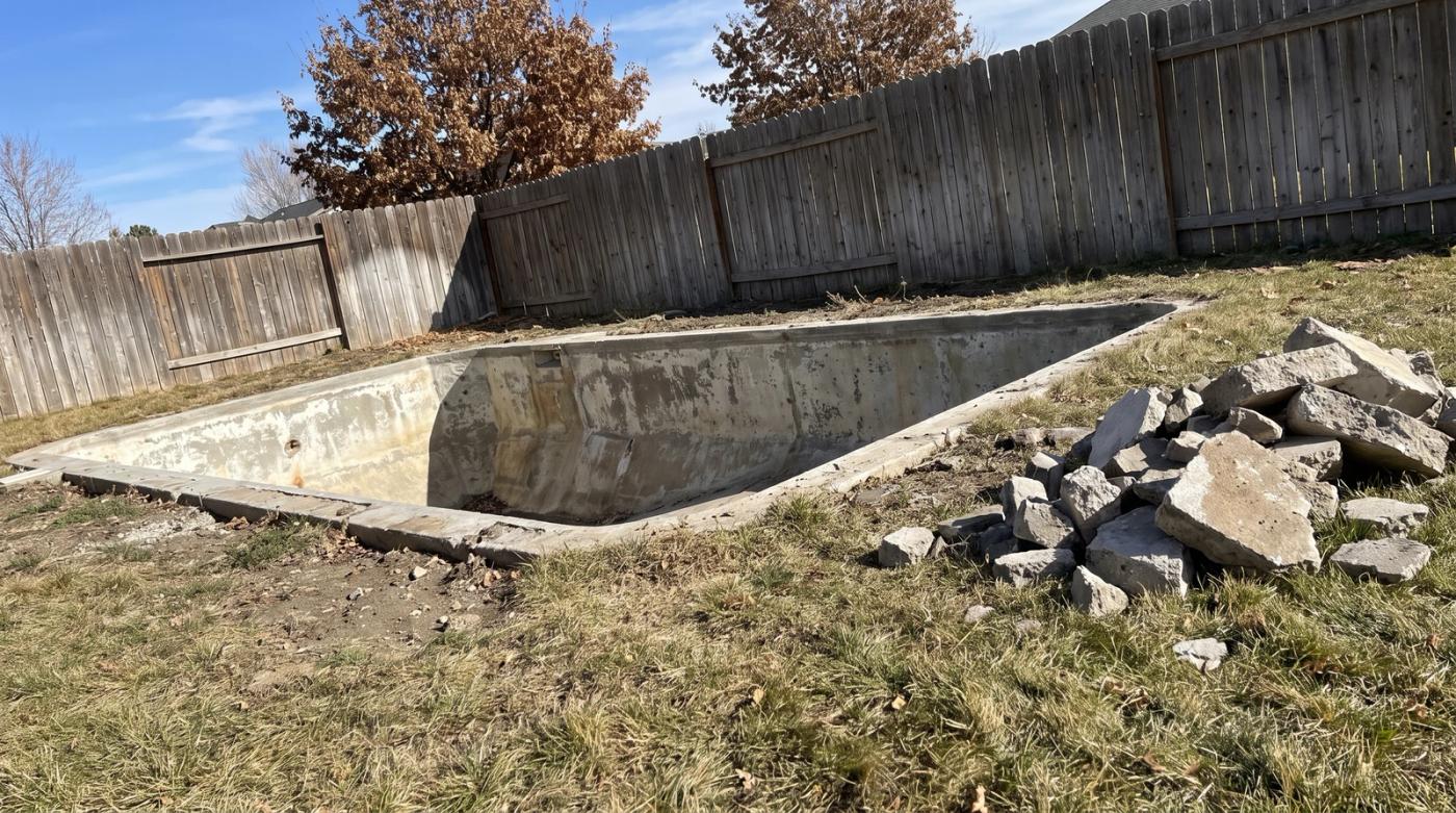 Empty concrete pool shell with broken concrete piled on the grass beside a wooden privacy fence during a pool demolition in Boise, Idaho