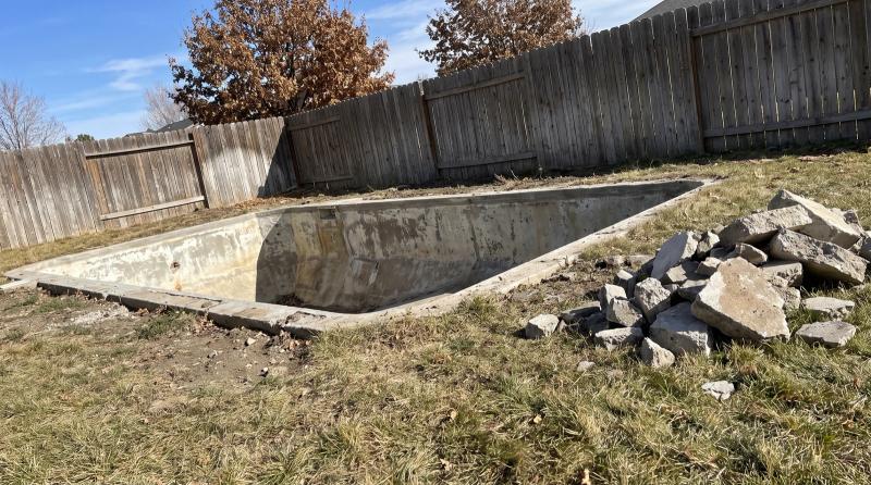 Empty concrete pool shell with broken concrete piled on the grass beside a wooden privacy fence during a pool demolition in Boise, Idaho