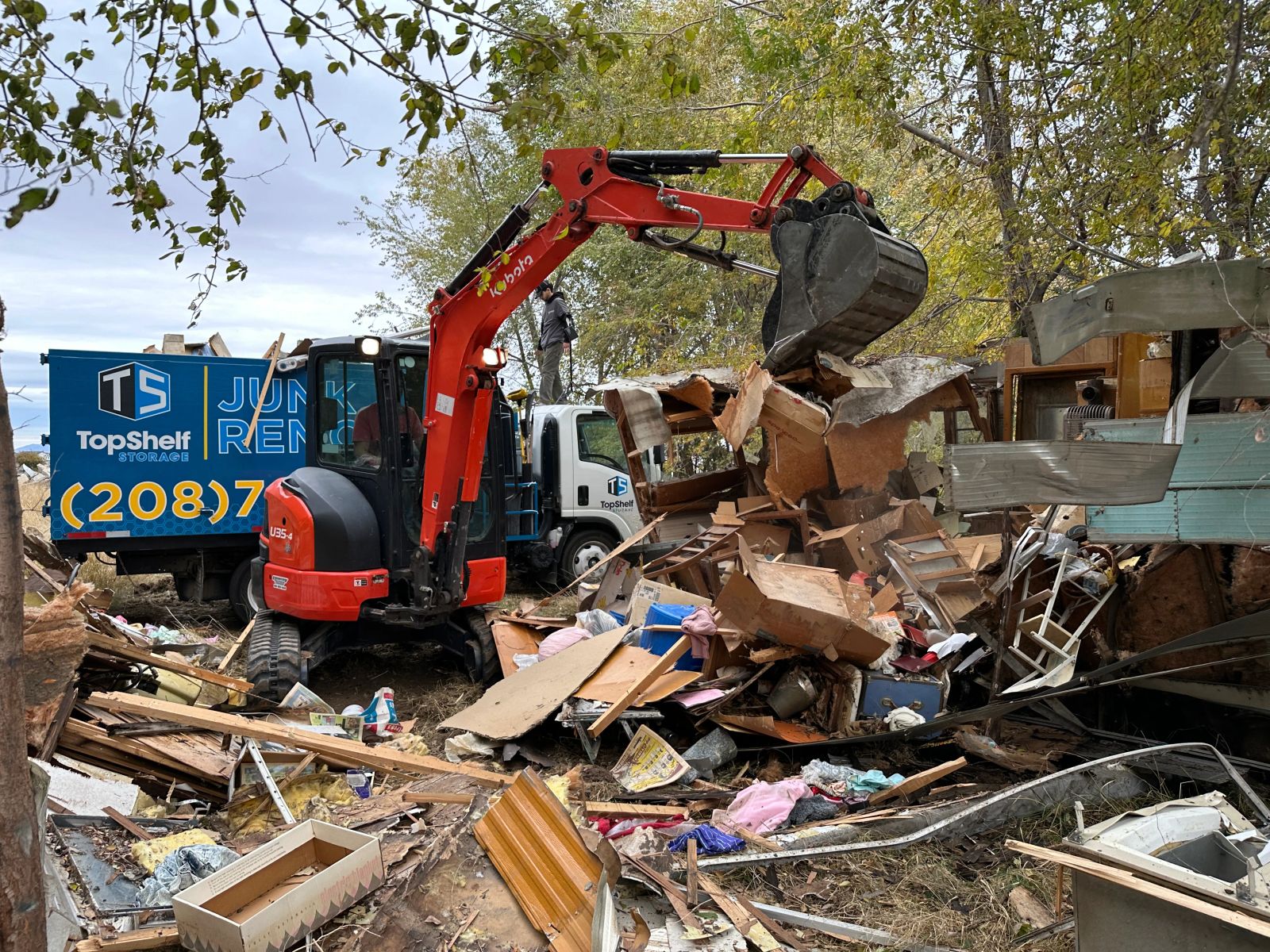 Top Shelf Kubota excavator demolishing an old RV with branded junk removal truck on a rural property in Nampa Idaho