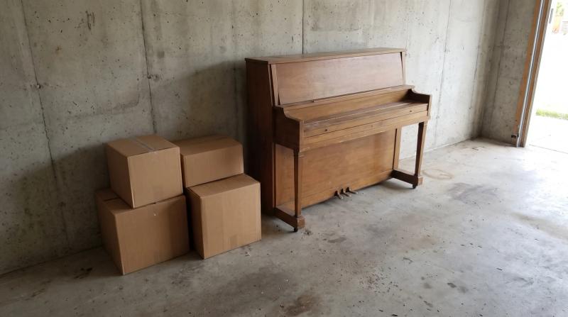 Old wooden upright piano on a garage floor next to stacked cardboard boxes, ready for junk removal pickup in Meridian, Idaho